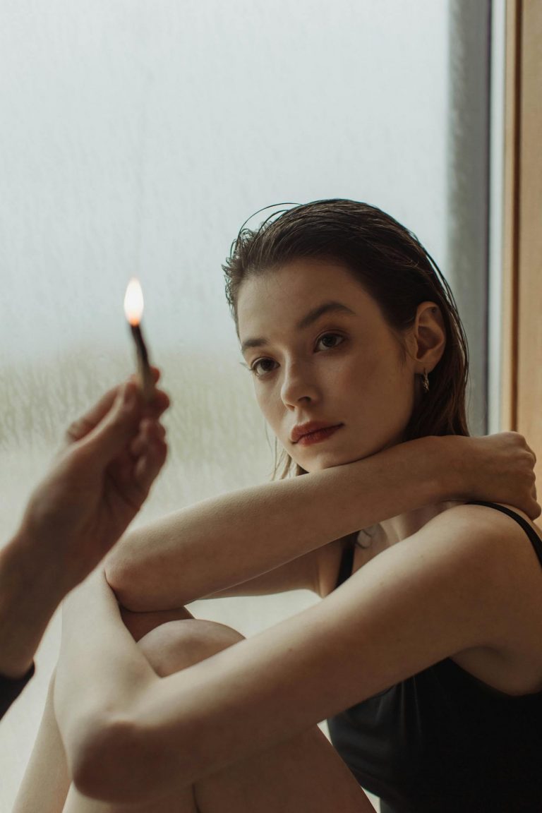 A woman sitting by a window, holding a palo santo stick, creating a reflective and serene mood.
